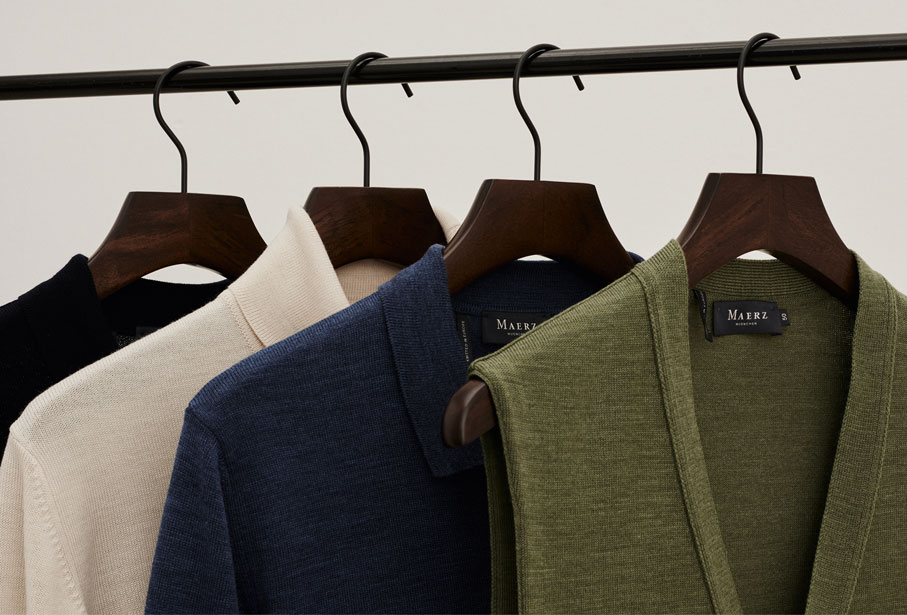 MAERZ Muenchen Merino Superwash Quality Detailbild/Nahaufnahme einer Kleiderstange mit vier Merino Superwash Artikel, einem Pullunder in grün, einem Polokragenpullover in blau, einem Rollkragenpullover in creme und einem Stehkragenpullover in schwarz.
