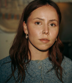 Frau mit rotbraunen Haaren in einer hellblauen, fein gestrickten Trachtenjacke mit Rundhalsausschnitt und Knopfleiste blickt in die Kamera