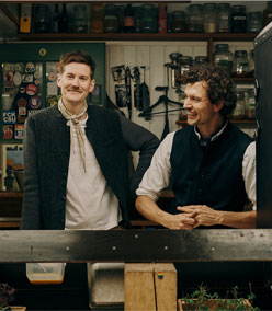 Lächelnde Männer in traditionell inspirierter, grauer Trachtenjacke mit Stehkragen und dunklen Knöpfen aus 100% Merinowolle, darunter ein weißes Shirt und Halstuch. Der andere daneben in einer dunkelblauer Weste mit Stehkragen und dunklen Knöpfen aus 100% Merinowolle, darunter ein weißes Hemd. Im Hintergrund Regale mit Gewürz- und Kräutergläsern.