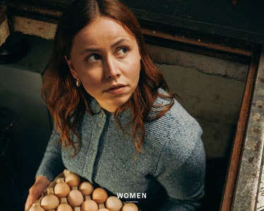 Frau mit rotbraunen Haaren in einer hellblauen, fein gestrickten Trachtenjacke mit Rundhalsausschnitt und Knopfleiste blickt in die Kamera.