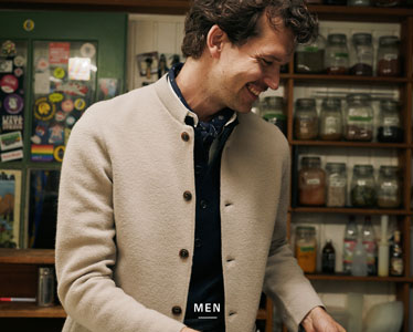 Lächelnder Mann in traditionell inspirierter, hellbeiger Trachtenjacke mit Stehkragen und dunklen Knöpfen aus 100% Merinowolle, darunter dunkelblaues Hemd und Halstuch. Im Hintergrund Regale mit Gewürz- und Kräutergläsern.