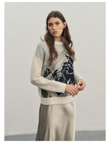 Eine Frau blickt in die Kamera und trägt einen weichen Pullover mit einem Bergmotiv in Pastelltönen sowie einen Rock in Creme.