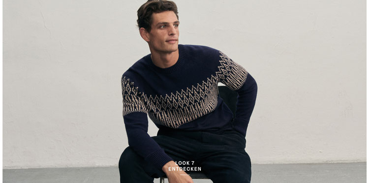 Mann in dunklem Strickpullover mit hellem Jacquard-Muster und passender Hose sitzt vor heller Wand.