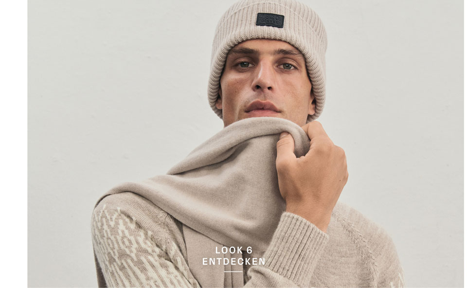 Ein Mann blickt in die Kamera und trägt eine beigefarbene Mütze, einen weichen Schal und einen Strickpullover in einem ausdrucksstarken Jacquard-Muster in Beige- und Cremetönen.