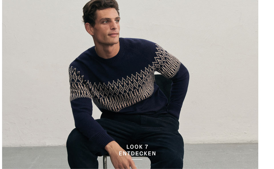Mann in dunklem Strickpullover mit hellem Jacquard-Muster und passender Hose sitzt vor heller Wand.
