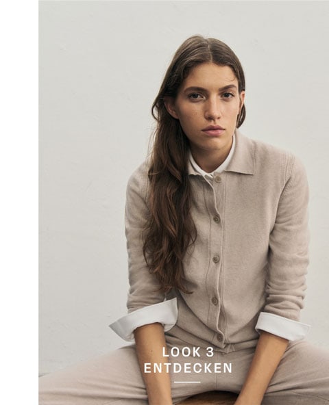 Favourite New Look Frau sitzt vor heller Wand in elegantem beigen Premium-Merino-Strickjacken-Outfit mit passender Hose und weißem Hemd.