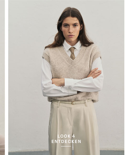Favourite New Look Eine Frau blickt in die Kamera, die Arme verschränkt. Sie trägt einen beigen Alpaka-Wollpullunder über einer weißen Bluse mit brauner Krawatte und eine Hose in Creme.