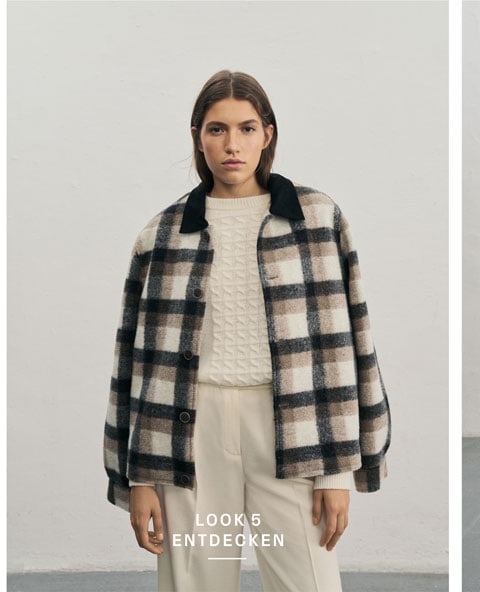 Favourite New Look Frau in lässigem Karo-Overshirt trägt weißen Pullover aus 100 % Schurwolle mit aufwendigem Strickmuster und passende Hose mit Bügelfalte, vor heller Wand.