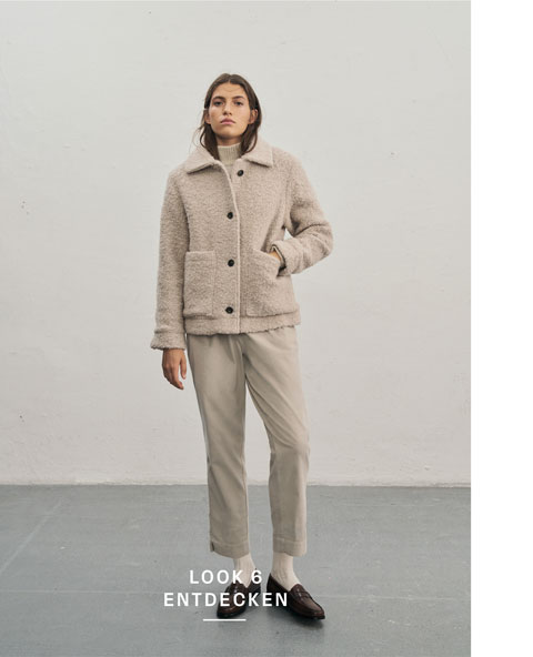 Favourite New Look Frau steht vor heller Wand in beiger Kuscheljacke im Teddylook, kombiniert mit Rollkragenpullover, passender Hose, hellen Socken und dunklen Lederschuhen.