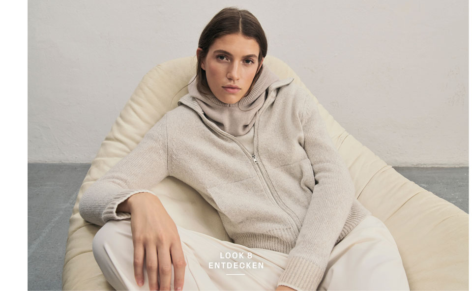 Favourite New Look Eine Frau sitzt entspannt in einem hellen Sessel und blickt in die Kamera. Sie trägt eine beigefarbene Strickjacke aus 100% Schurwolle und eine dazu passende Balaklava, kombiniert mit einer hellen Hose.