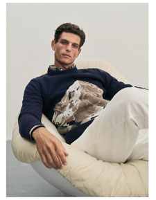 Mann sitzt lässig auf einem Sessel und trägt einen weichen Pullover mit einem Bergmotiv sowie eine weiße Hose.