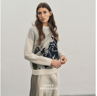 Favourite New Look Eine Frau blickt in die Kamera und trägt einen weichen Pullover mit einem Bergmotiv in Pastelltönen sowie einen Rock in Creme.