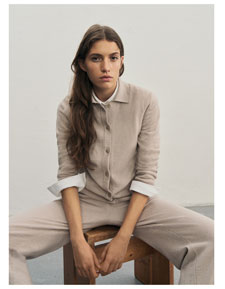 Frau sitzt vor heller Wand in elegantem beigen Premium-Merino-Strickjacken-Outfit mit passender Hose und weißem Hemd.