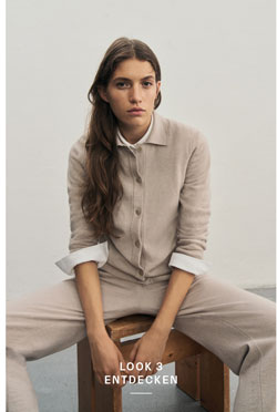 Frau in elegantem beigen Premium-Merino-Strickjacken-Outfit mit passender Hose und weißem Hemd vor heller Wand.