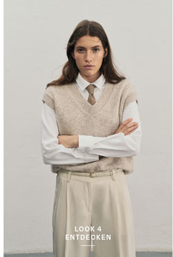 Eine Frau blickt in die Kamera, die Arme verschränkt. Sie trägt einen beigen Alpaka-Wollpullunder über einer weißen Bluse mit brauner Krawatte und eine Hose in Creme.