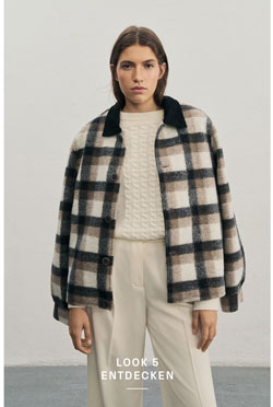 Frau in lässigem Karo-Overshirt trägt weißen Pullover aus 100 % Schurwolle mit aufwendigem Strickmuster und passende Hose mit Bügelfalte, vor heller Wand.