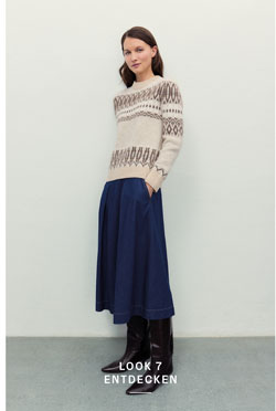 Frau in beigem Pullover mit dunklem Strickmuster aus 100 % Schurwolle, blauem Jeansrock und braunen Lederstiefeln vor heller Wand.