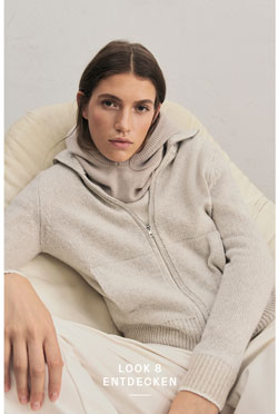 Eine Frau sitzt entspannt in einem hellen Sessel und blickt in die Kamera. Sie trägt eine beigefarbene Strickjacke aus 100% Schurwolle und eine dazu passende Balaklava, kombiniert mit einer hellen Hose.