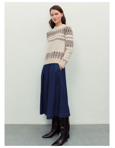 Frau vor heller Wand in beigem Strickpullover mit dunklem Jacquard-Muster aus 100 % Schurwolle, blauem Jeansrock und braunen Lederstiefeln.