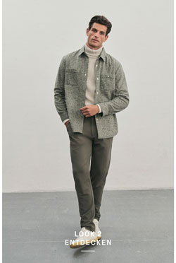 Mann steht vor heller Wand in hellem Strick-Rollkragenpullover mit dunkler Hose und weißen Sneakern, kombiniert mit markantem Fischgrät-Overshirt.