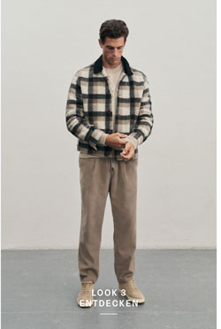 Mann steht vor heller Wand in beigem Outfit aus hellem Strickpullover, beiger Hose und Sneakern, kombiniert mit markantem Karo-Overshirt in Navy, Beige und Weiß.