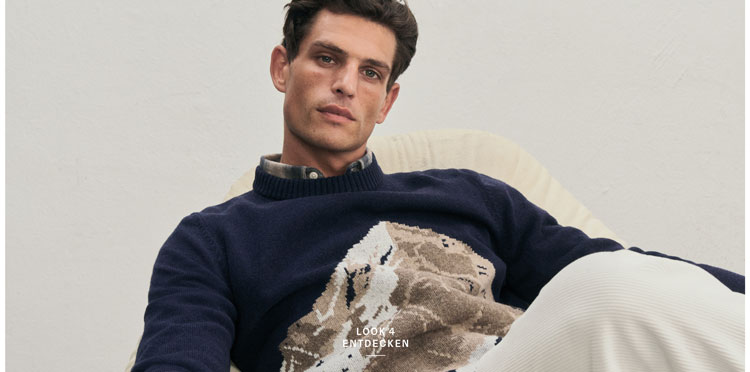 Mann sitzt lässig auf einem Sessel und trägt einen weichen Pullover mit einem Bergmotiv sowie eine weiße Hose.
