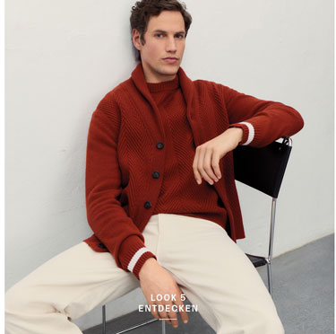 Mann sitzt vor heller Wand in eleganter dunkelroter Strick-Kombination aus Strickjacke mit weißen Ärmelnstreifen, Pullover und beiger Hose.