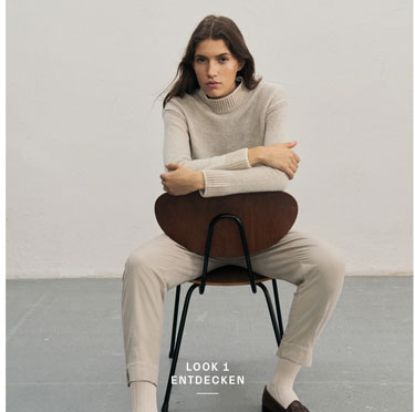 Favourite New Look Frau in elegantem beigen Ton-in-Ton-Outfit mit Stehkragenpullover, Hose und Socken sowie braunen Schuhen vor heller Wand.