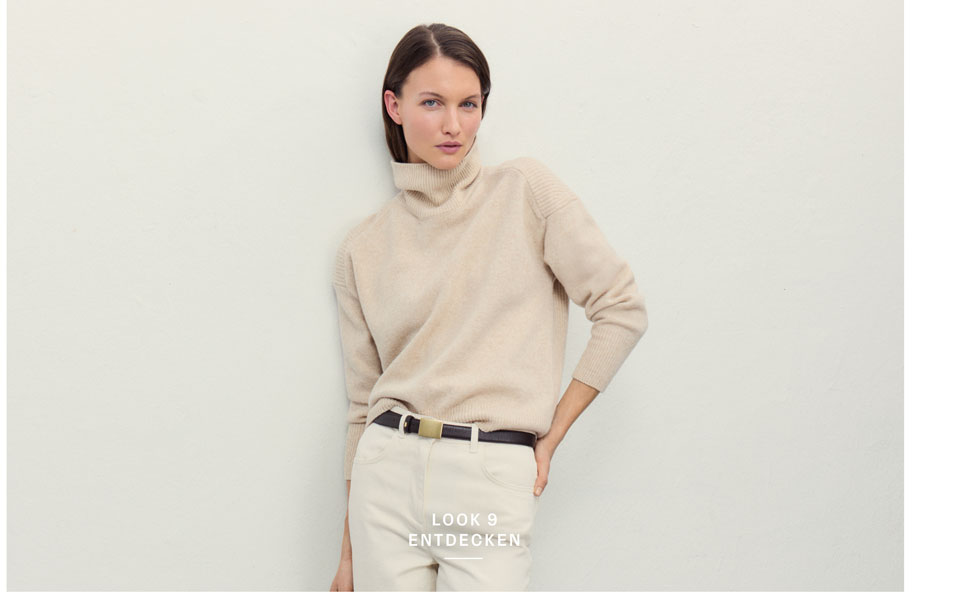 Favourite New Look Eine Frau steht vor einer hellen Wand und blickt ruhig in die Kamera. Sie trägt einen beigen Rollkragenpullover aus 100% Schurwolle, eine weiße Hose mit Gürtel.