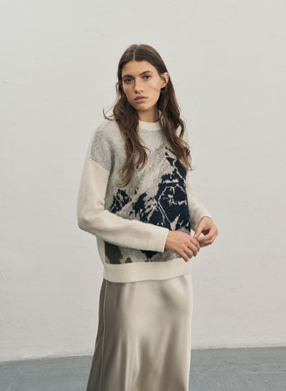Eine Frau blickt in die Kamera und trägt einen weichen Pullover mit einem Bergmotiv in Pastelltönen sowie einen Rock in Creme.