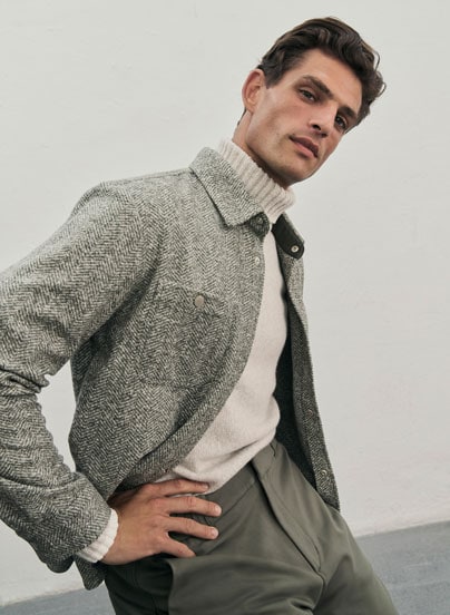 Mann vor neutralem Hintergrund in hellem Merinowoll-Rollkragenpullover, dunkler Hose und Fischgrät-Overshirt.