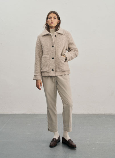 Frau steht vor heller Wand in beiger Kuscheljacke im Teddylook, kombiniert mit Rollkragenpullover, passender Hose, hellen Socken und dunklen Lederschuhen.