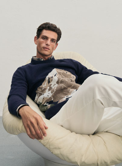 Mann sitzt lässig auf einem Sessel und trägt einen weichen Pullover mit einem Bergmotiv sowie eine weiße Hose.