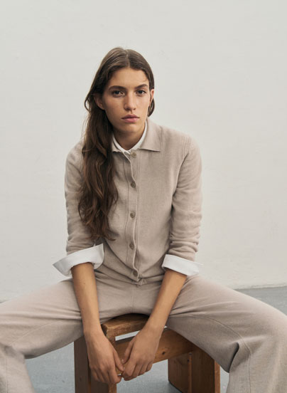 Frau sitzt vor heller Wand in elegantem beigen Premium-Merino-Strickjacken-Outfit mit passender Hose und weißem Hemd.