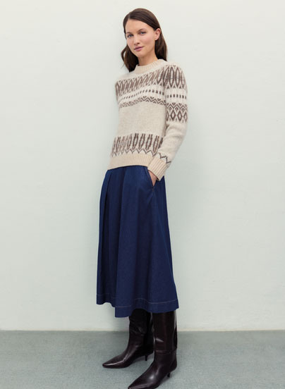 Frau vor heller Wand in beigem Strickpullover mit dunklem Jacquard-Muster aus 100 % Schurwolle, blauem Jeansrock und braunen Lederstiefeln.