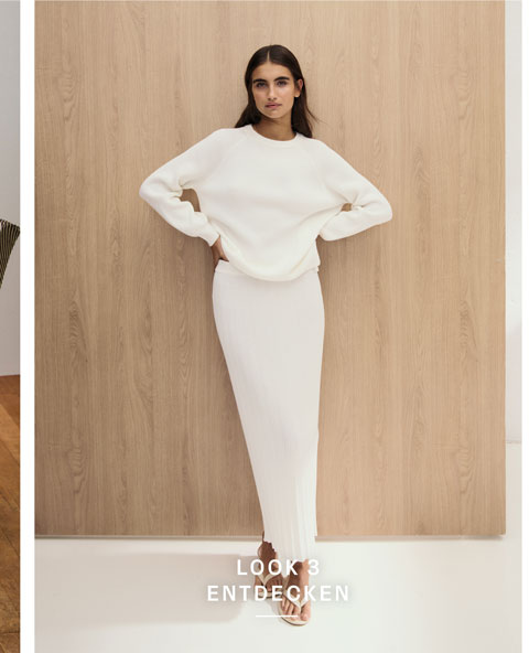 Look 3 entdecken: Our Favourite New Looks. Frau im hellen Strick Set aus Pullover und Rock, minimalistisch vor Holzpaneel inszeniert.