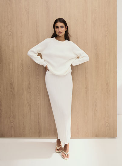 Our Favourite New Looks: Frau im hellen Strick Set aus Pullover und Rock, minimalistisch vor Holzpaneel inszeniert.