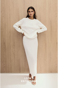 Look 3 entdecken: Our Favourite New Looks. Frau im hellen Strick Set aus Pullover und Rock, minimalistisch vor Holzpaneel inszeniert.