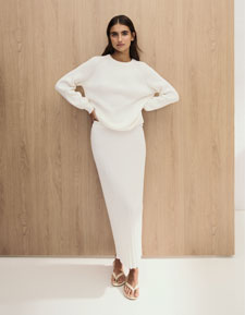 Our Favourite New Looks: Frau im hellen Strick Set aus Pullover und Rock, minimalistisch vor Holzpaneel inszeniert.