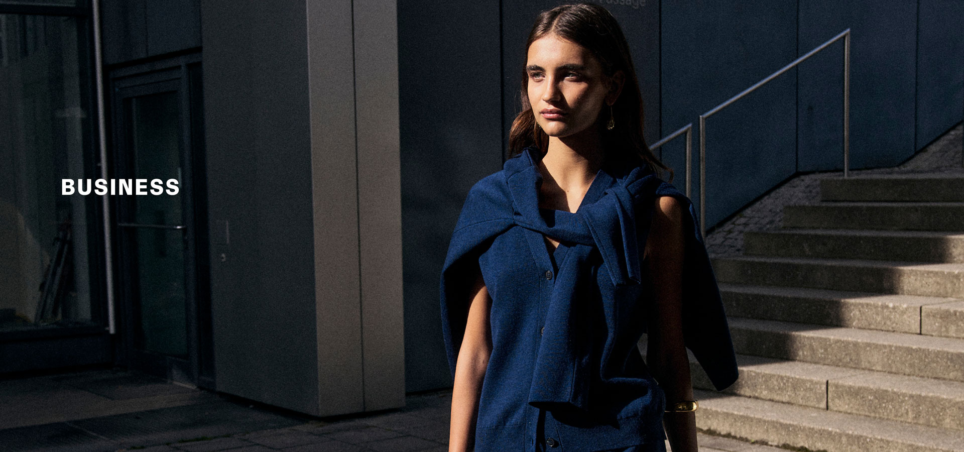 Frau im dunkelblauen BUSINESS Look mit locker umgelegtem Strick und Pullunder, urban vor moderner Fassade inszeniert – Headline ‚BUSINESS‘.