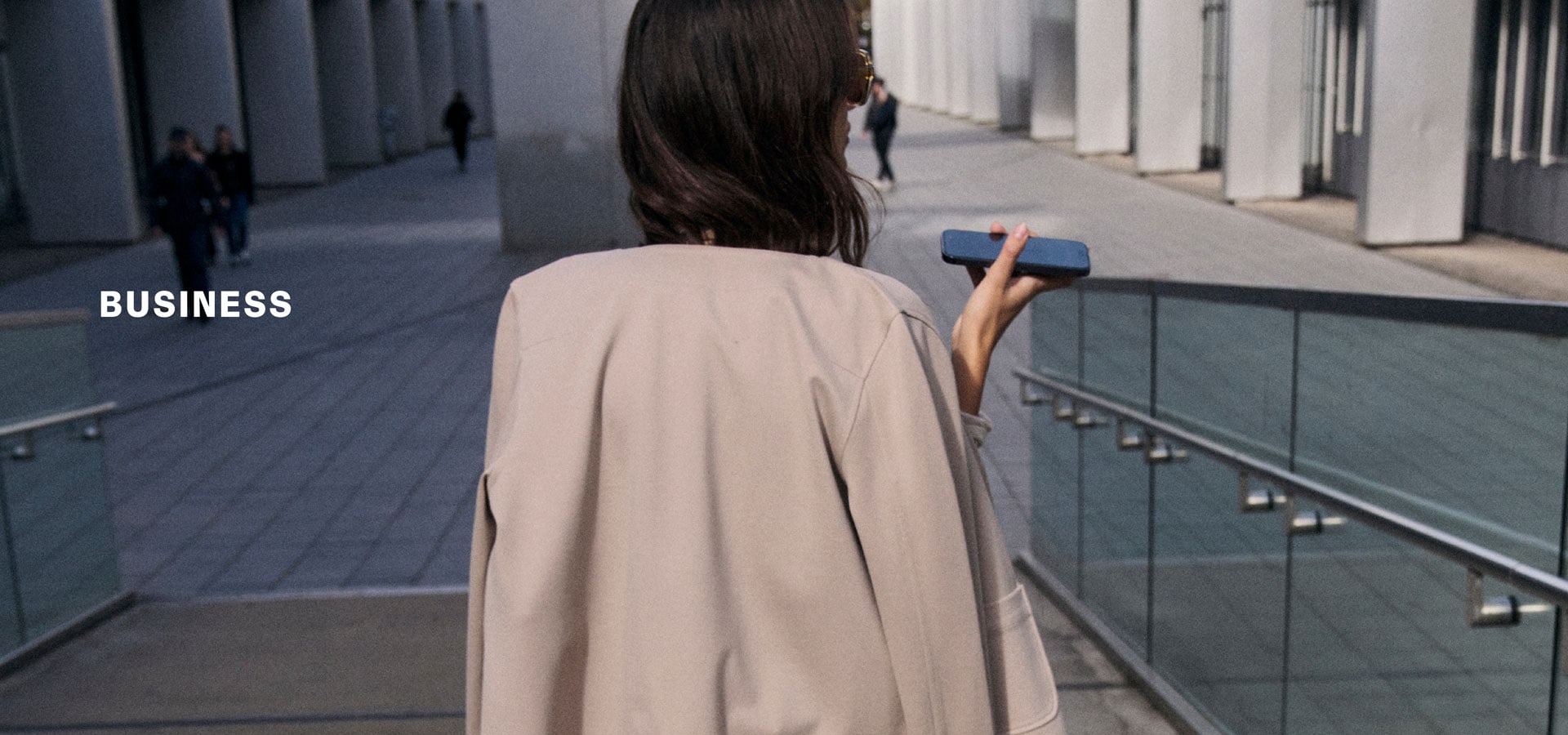 Rückenansicht einer Frau in hellem Business‑Look mit fließender Jacke und Hose, auf urbaner Passage unterwegs – inszeniert neben der Headline ‚BUSINESS‘.