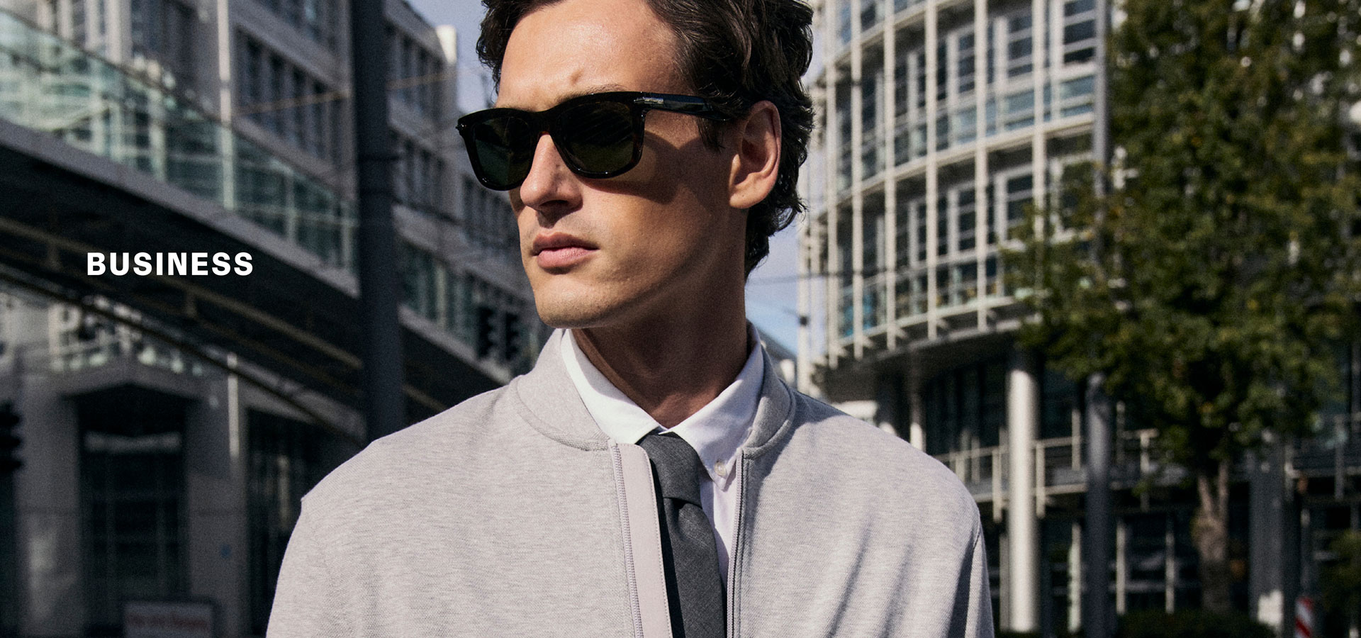 Mann mit Sonnenbrille im hellen Business‑Look mit Jacke, Hemd und Krawatte, urban vor moderner Architektur inszeniert – neben der Headline ‚BUSINESS‘.
