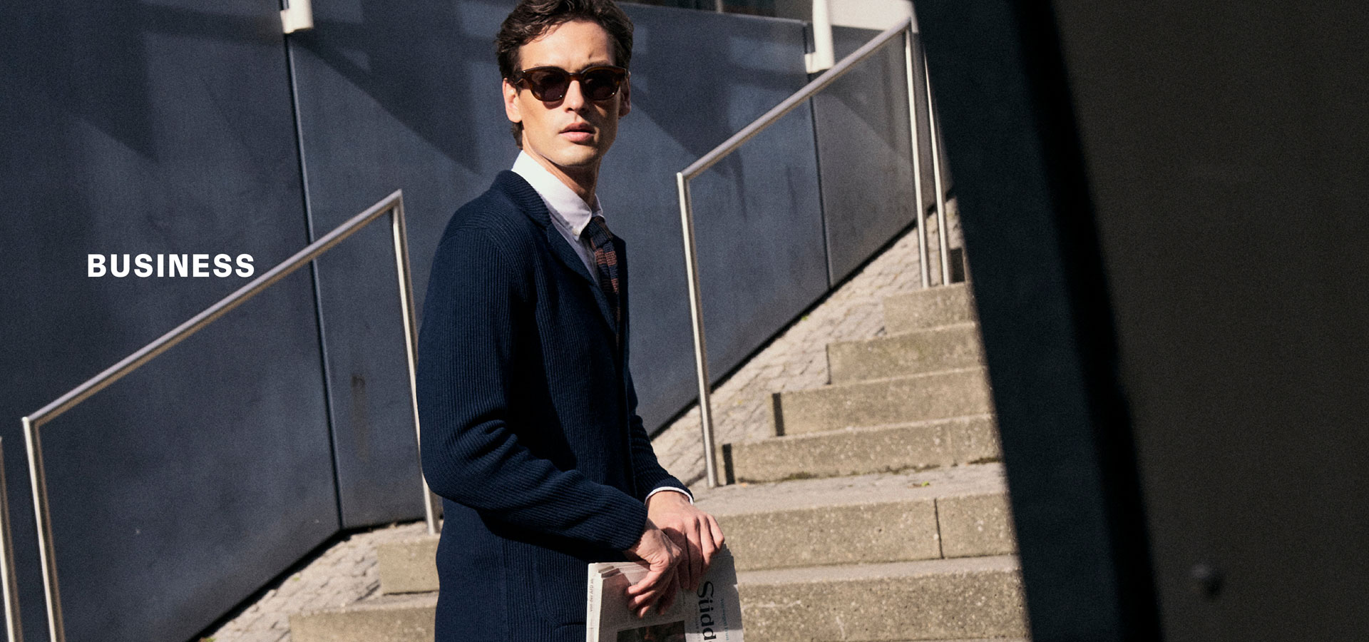Mann mit Sonnenbrille in dunkelblauem Business‑Look mit Strickjacke und Hemd auf Treppe urban inszeniert – neben der Headline ‚BUSINESS‘.