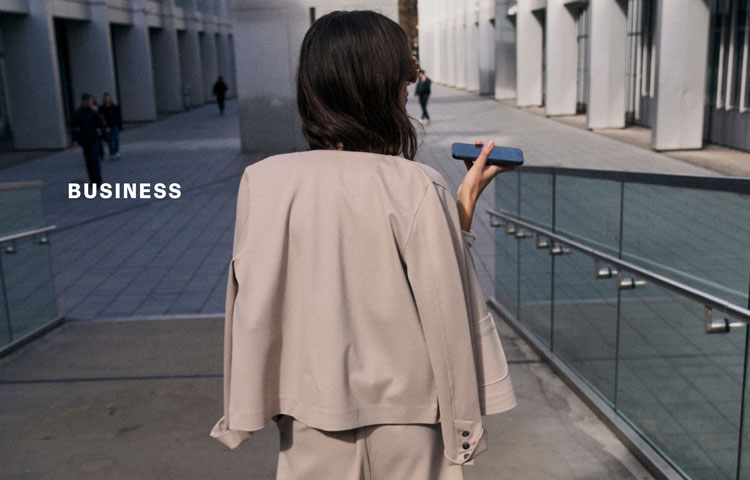 Rückenansicht einer Frau in hellem Business‑Look mit fließender Jacke und Hose, auf urbaner Passage unterwegs – inszeniert neben der Headline ‚BUSINESS‘.