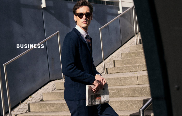 Mann mit Sonnenbrille in dunkelblauem Business‑Look mit Strickjacke und Hemd auf Treppe urban inszeniert – neben der Headline ‚BUSINESS‘.
