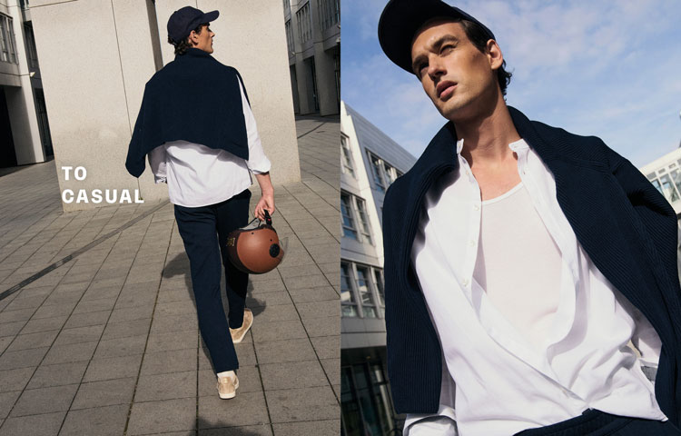 Mann in lässigem TO CASUAL Look mit dunkelblauem Strick, weißem Shirt und Cap, urban inszeniert vor moderner Architektur.