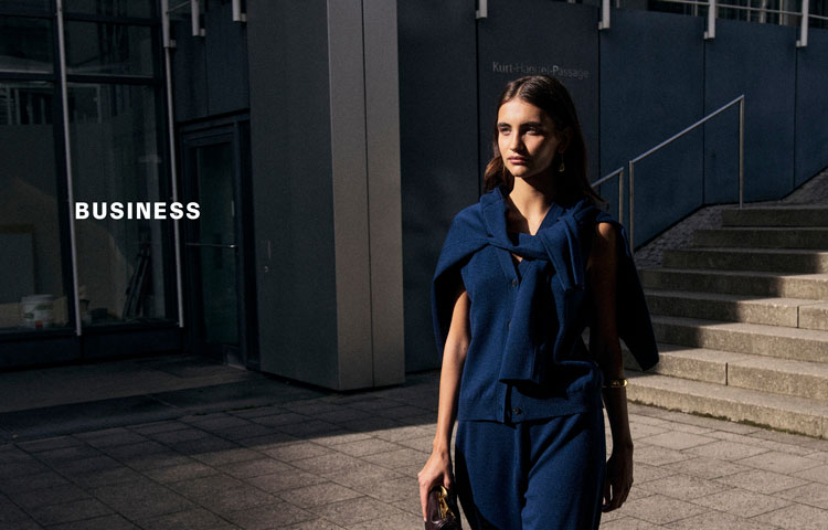 Frau im dunkelblauen BUSINESS Look mit locker umgelegtem Strick und Pullunder, urban vor moderner Fassade inszeniert – Headline ‚BUSINESS‘.