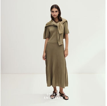 Look 12 entdecken: Our Favourite New Looks. Frau in olivfarbenem Kleid und umgelegter Strickjacke, minimalistisch im Studio inszeniert.