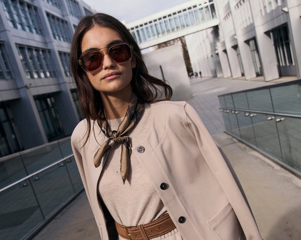 Monday to Friday: Frau im hellen Layer‑Look mit Blazer, Gürtel und Sonnenbrille, modern im urbanen Außenraum inszeniert.