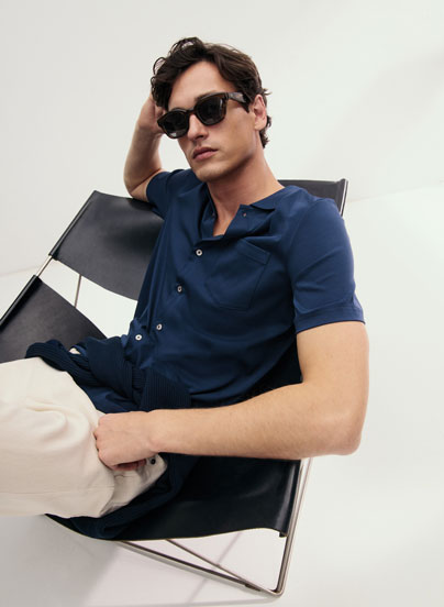 Look 1 entdecken: Our Favourite New Looks: Mann im navyfarbenen Stricktop und heller Hose, locker im Designerstuhl positioniert, minimalistisches Studio Setting.